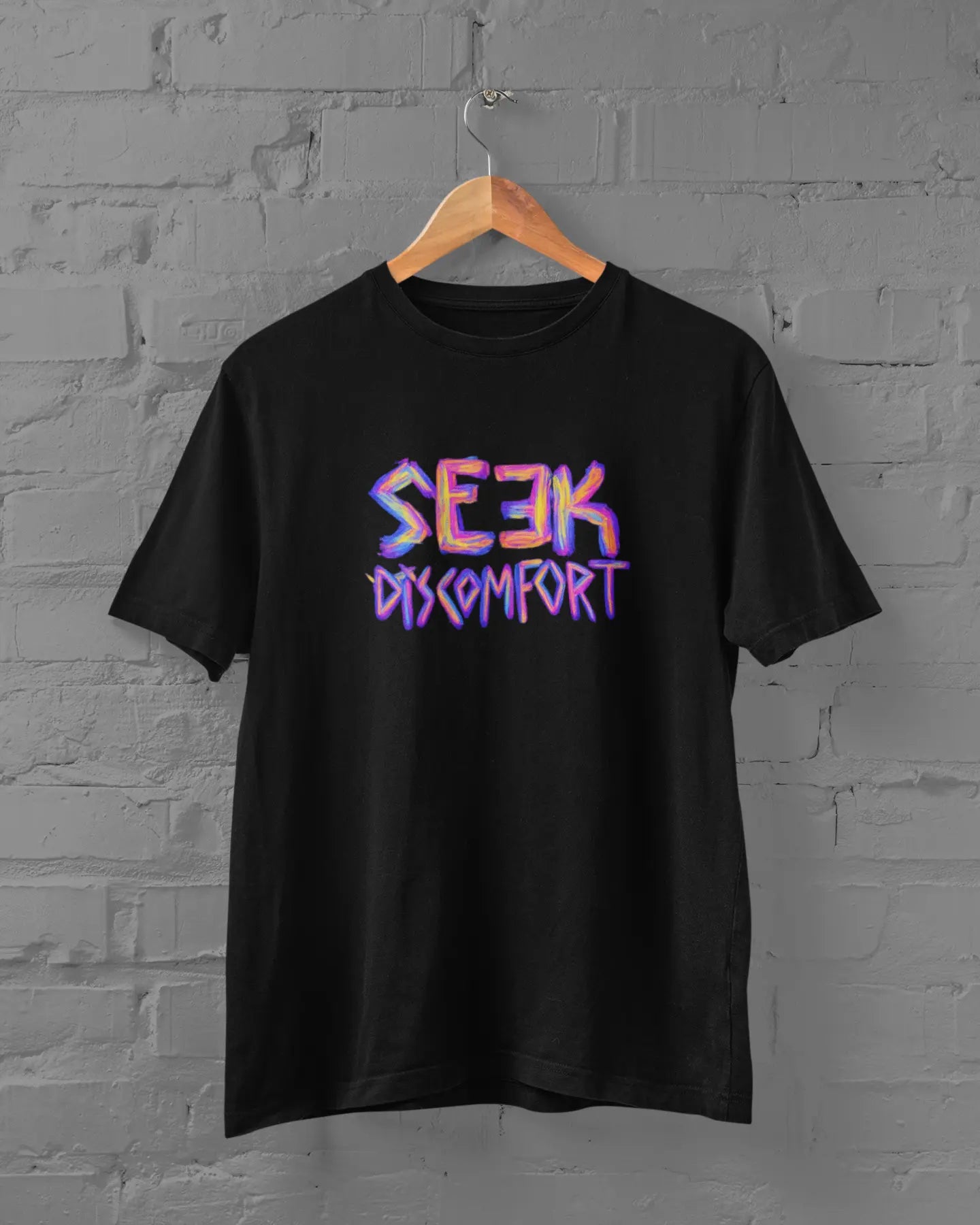 Black t-shirt with 'Seek Discomfort' text on a hanger against a gray brick wall.