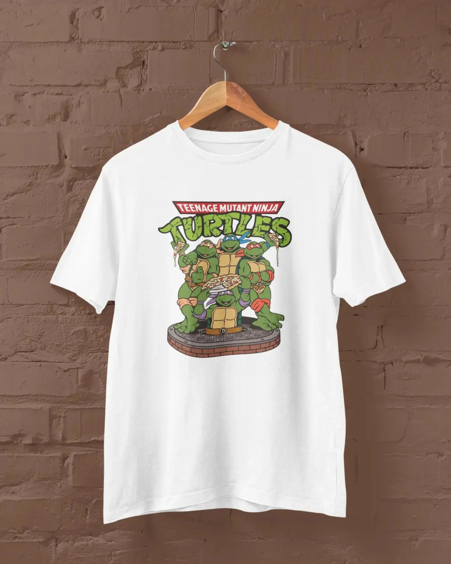White t-shirt with Teenage Mutant Ninja Turtles graphic on a hanger against a brown brick wall.