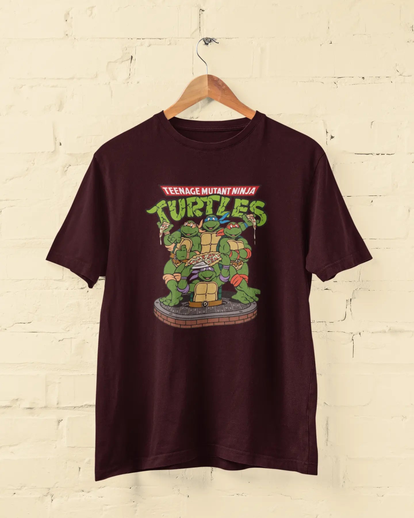 Maroon t-shirt with Teenage Mutant Ninja Turtles graphic on a hanger against a beige brick wall.