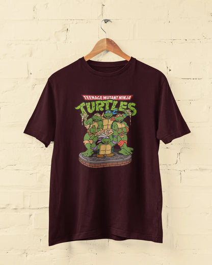 Maroon t-shirt with Teenage Mutant Ninja Turtles graphic on a hanger against a beige brick wall.