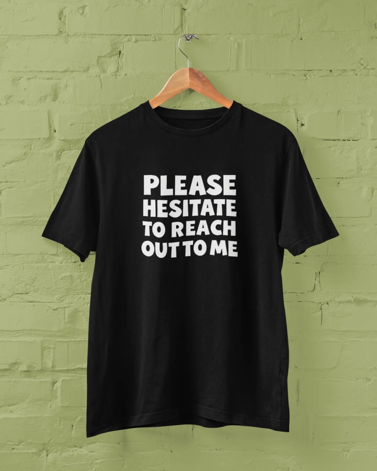 Black t-shirt with 'PLEASE HESITATE TO REACH OUT TO ME' text on a green brick wall background